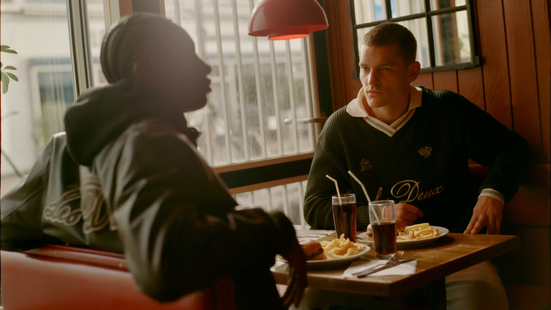 Two men sitting across from each other in a diner booth, sharing a plate of fries and a drink. The man on the right wears a dark green V-neck soccer sweater with 'Les Deux' embroidered on the chest, while the man on the left wears a dark hoodie.