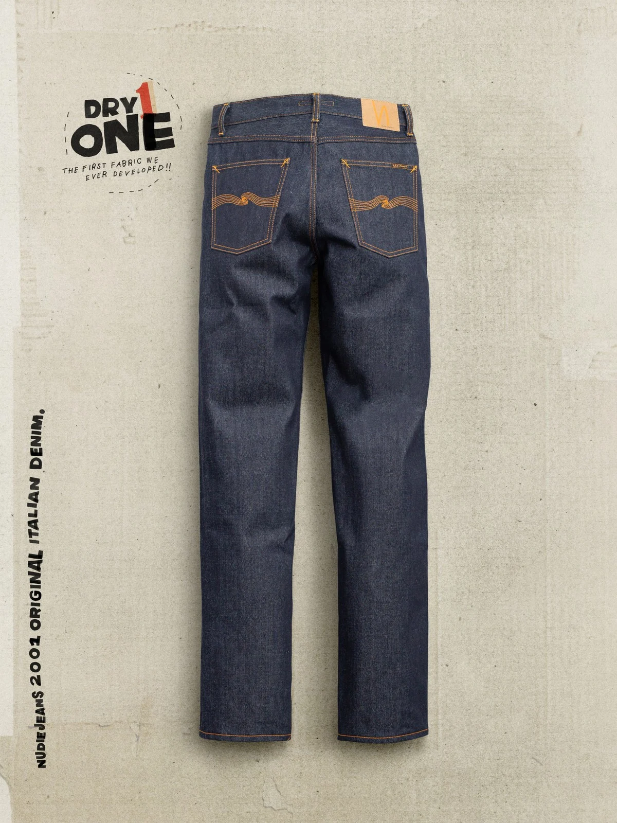 A pair of dark blue denim jeans laid flat backwards against a textured light beige background, with branding text 'DRY ONE' and 'NUDIE JEANS 2001 ORIGINAL ITALIAN DENIM' printed on the left side.