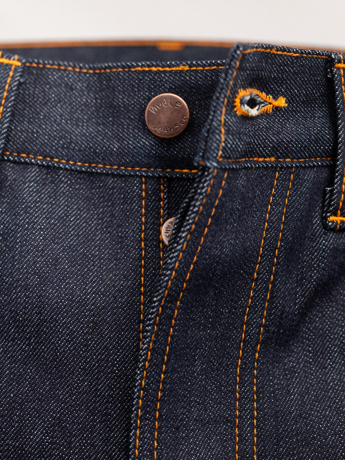 Close-up of dark indigo denim fabric with bright orange contrast stitching and copper-toned metal buttons.