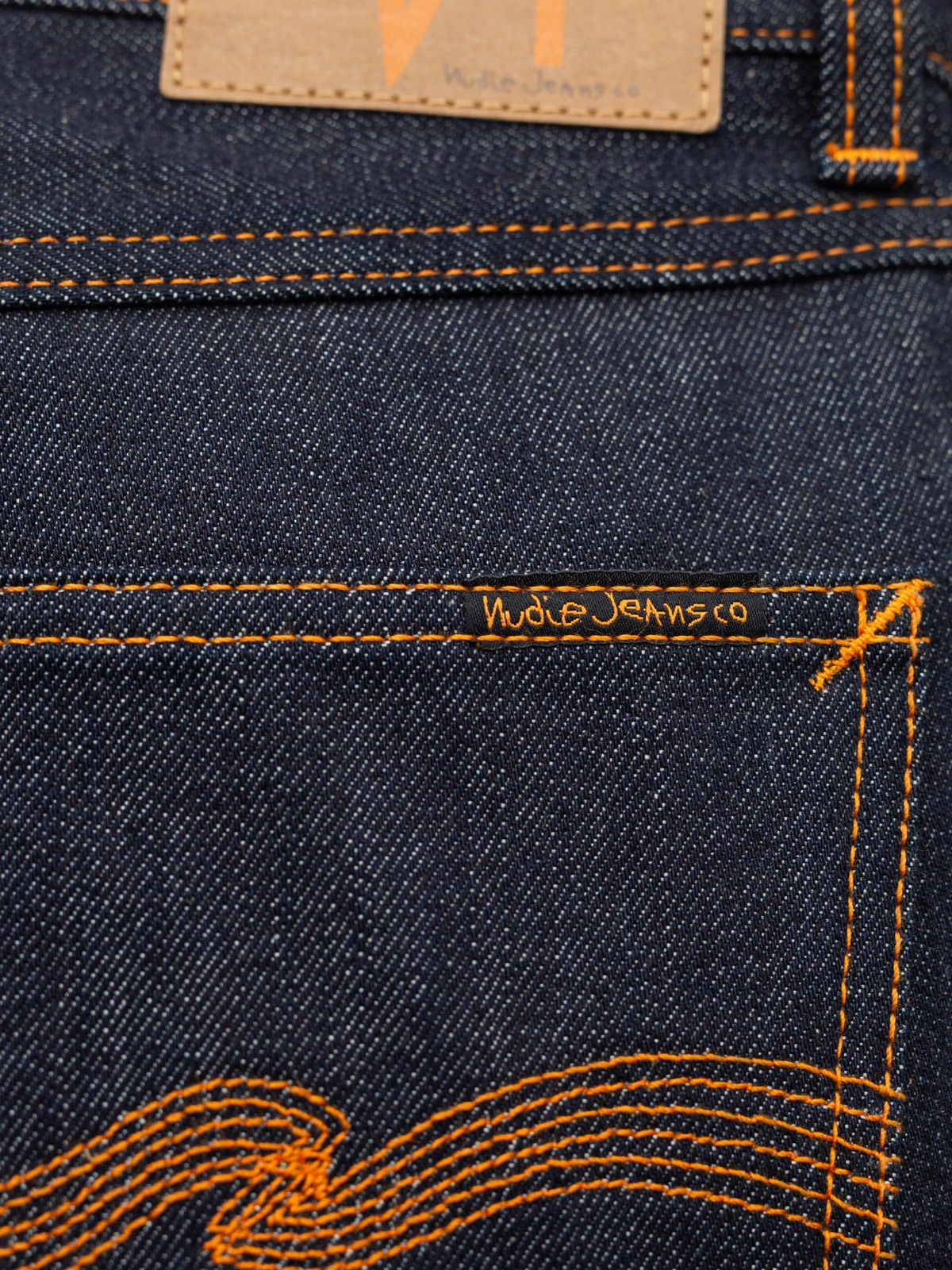Close-up of dark indigo denim jeans showing orange contrast stitching, a tan leather patch, and the embroidered 'Nudie Jeans co' brand name on the back pocket.